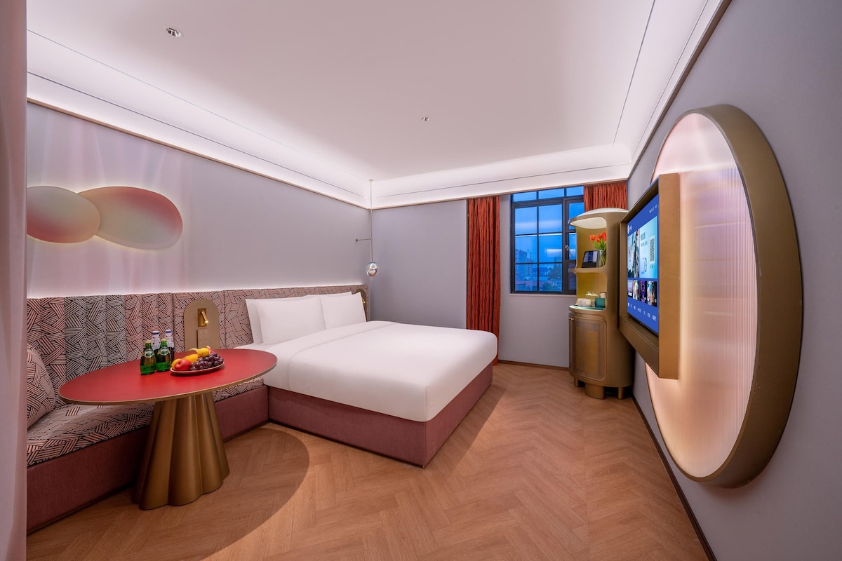 Une chambre d’hôtel moderne équipée d’un lit, d’un coin salon, d’une télévision et d’une petite table avec des boissons et des fruits.