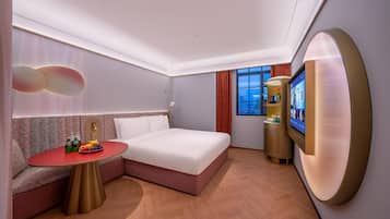 A modern hotel room with a bed, a seating area, a TV, and a small table with drinks and fruit.