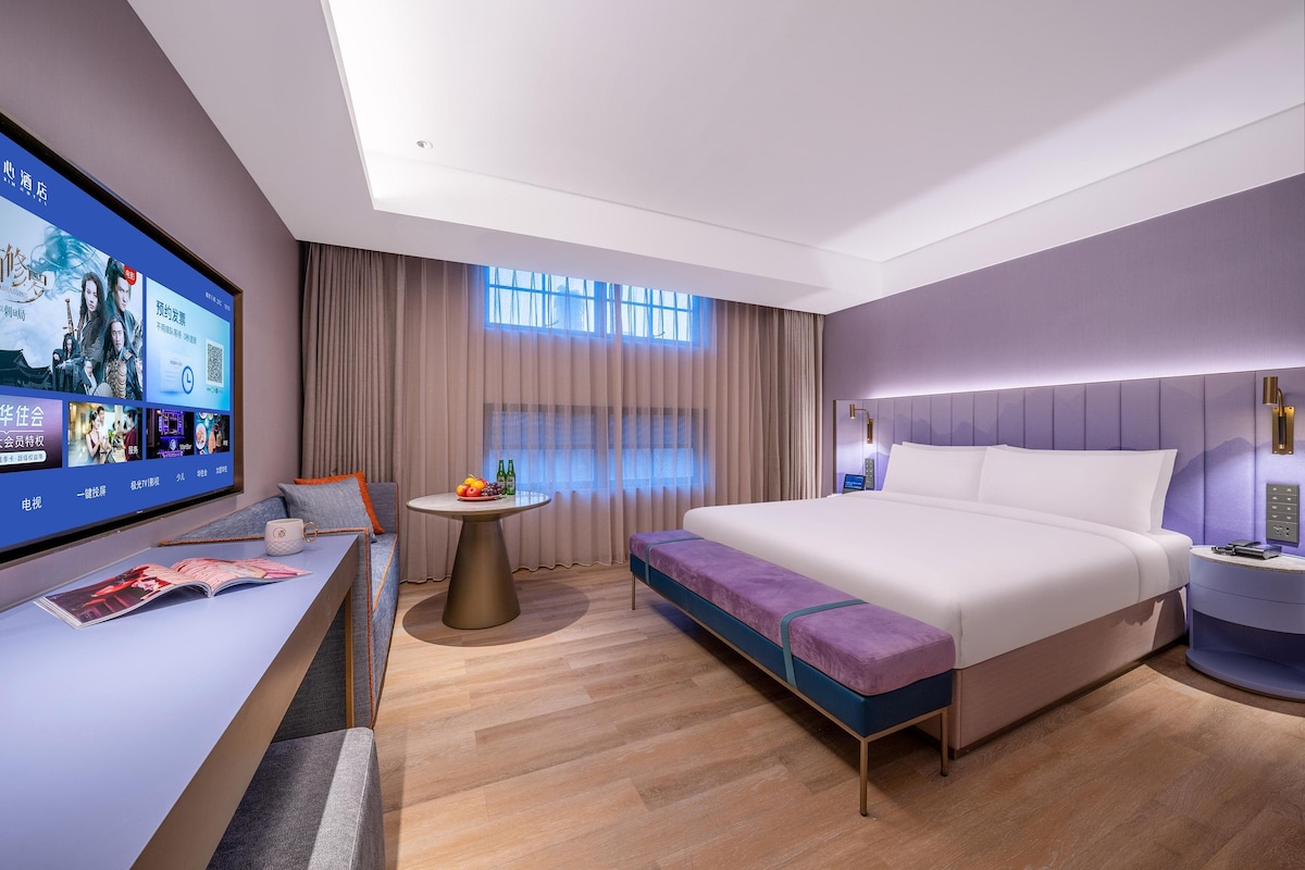Une chambre d’hôtel moderne dotée d’un grand téléviseur à écran plat, d’un canapé, d’une petite table ronde avec des fruits, d’un lit agrémenté de touches de couleur violette et d’une table de chevet avec une lampe.