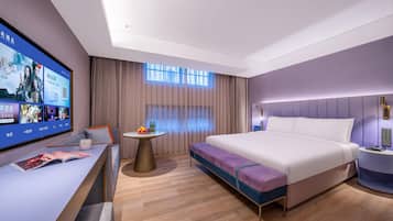 A modern hotel room with a large flat-screen TV, a sofa, a small round table with fruits, a bed with a purple accent, and a nightstand with a lamp.
