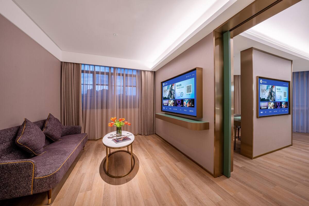 Ein modernes Hotelzimmer mit einer Couch, einem kleinen Tisch mit einer Vase und zwei großen digitalen Bildschirmen, die Inhalte zeigen.