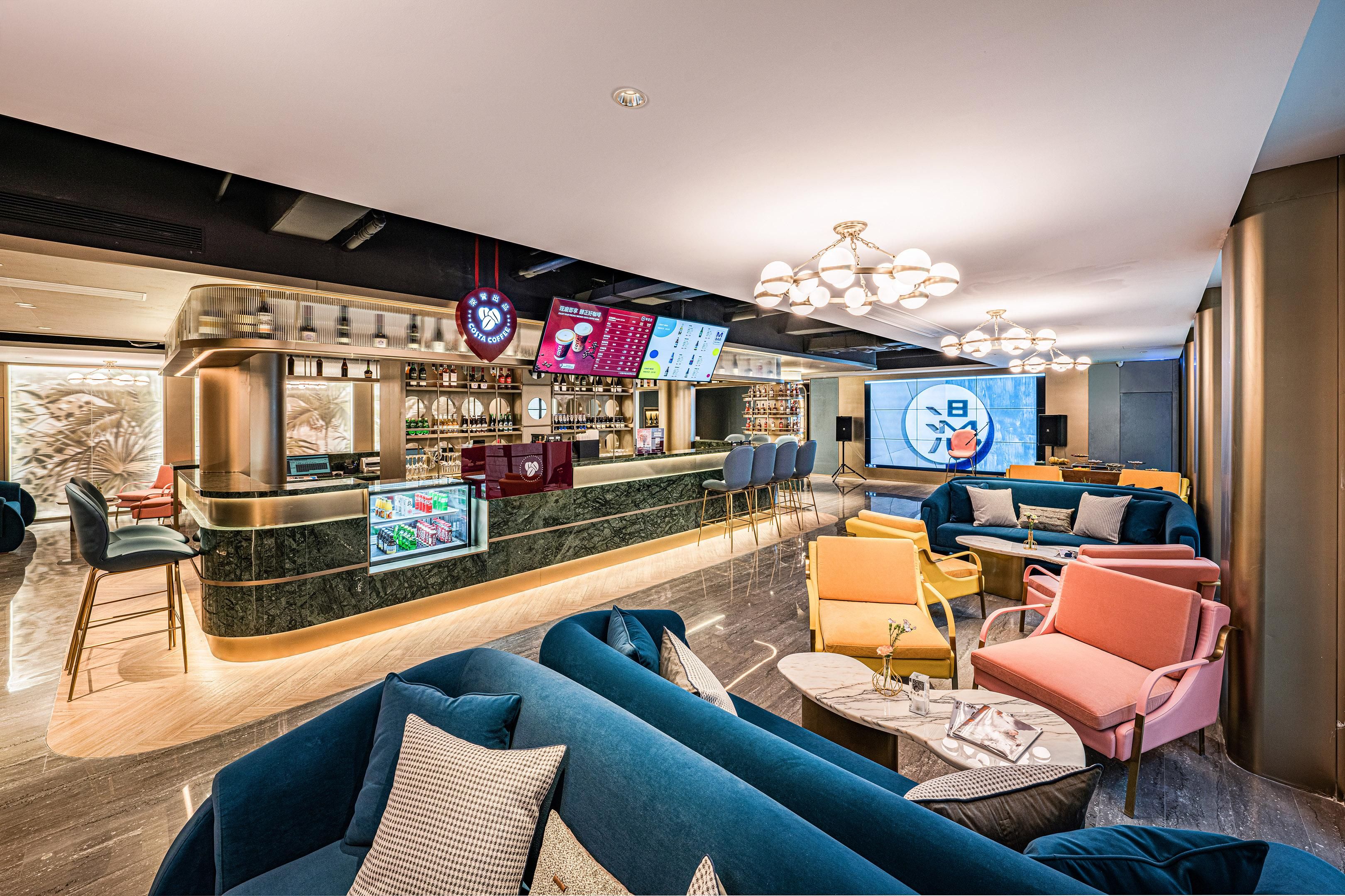Zona de bar moderna con barra de mármol, asientos con sofás azul y rosa, y una pantalla grande mostrando contenido.