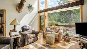 A cozy living room with leather sofas, a wooden coffee table, and a mounted deer head. Large windows offer a view of the outdoors. - Elk River Cabin -- Cabin Condo In Estes Park -- 23-zone3554 3 Bedroom Condo by RedAwning (Estes Park)