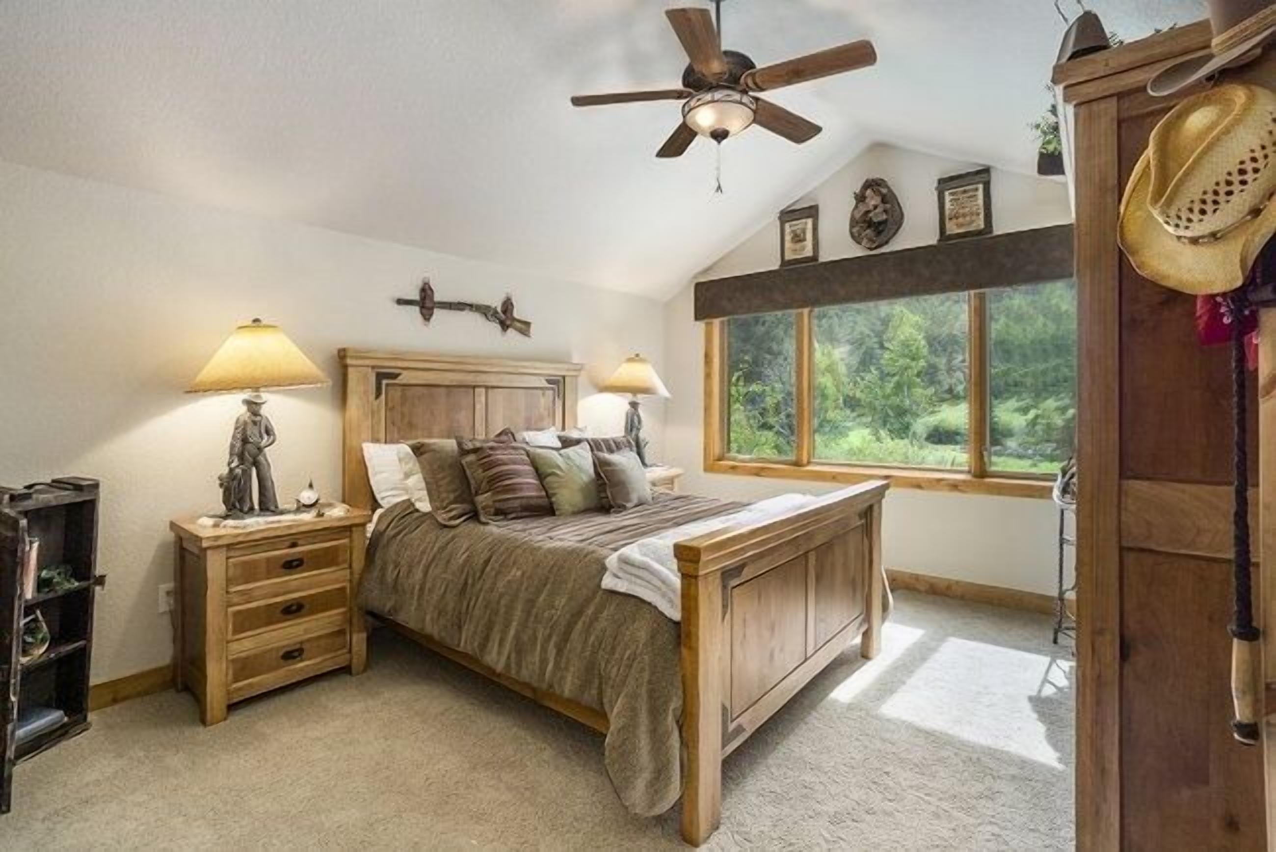 Quarto com cama de madeira, duas mesas de cabeceira com luminárias, ventilador de teto, janela grande com vista para vegetação e guarda-roupa de madeira.