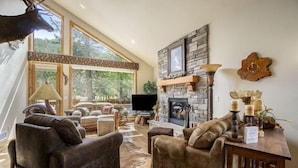 A spacious living room with leather sofas, a stone fireplace, a mounted deer head, and a large window overlooking the outdoors. - Elk River Cabin -- Cabin Condo In Estes Park -- 23-zone3554 3 Bedroom Condo by RedAwning (Estes Park)