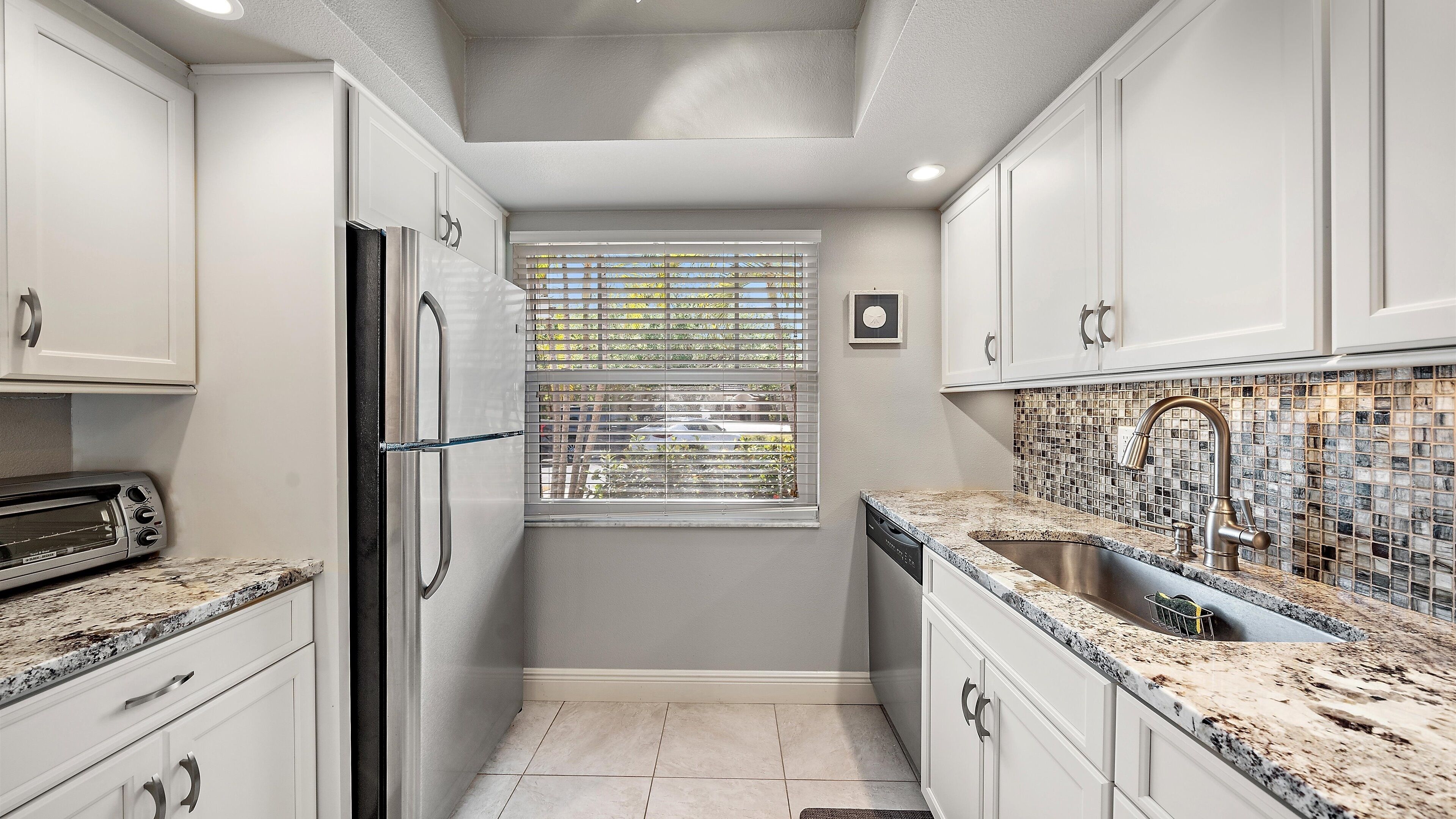Cocina compacta con armarios blancos, horno empotrado, ventana con persianas y salpicadero de azulejos.