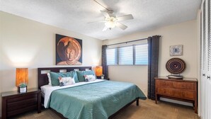 A bedroom with a bed, nightstands, a ceiling fan, a window with curtains, and a wooden dresser. - Island Surf 614 2 Bedroom Apts (Kihei)