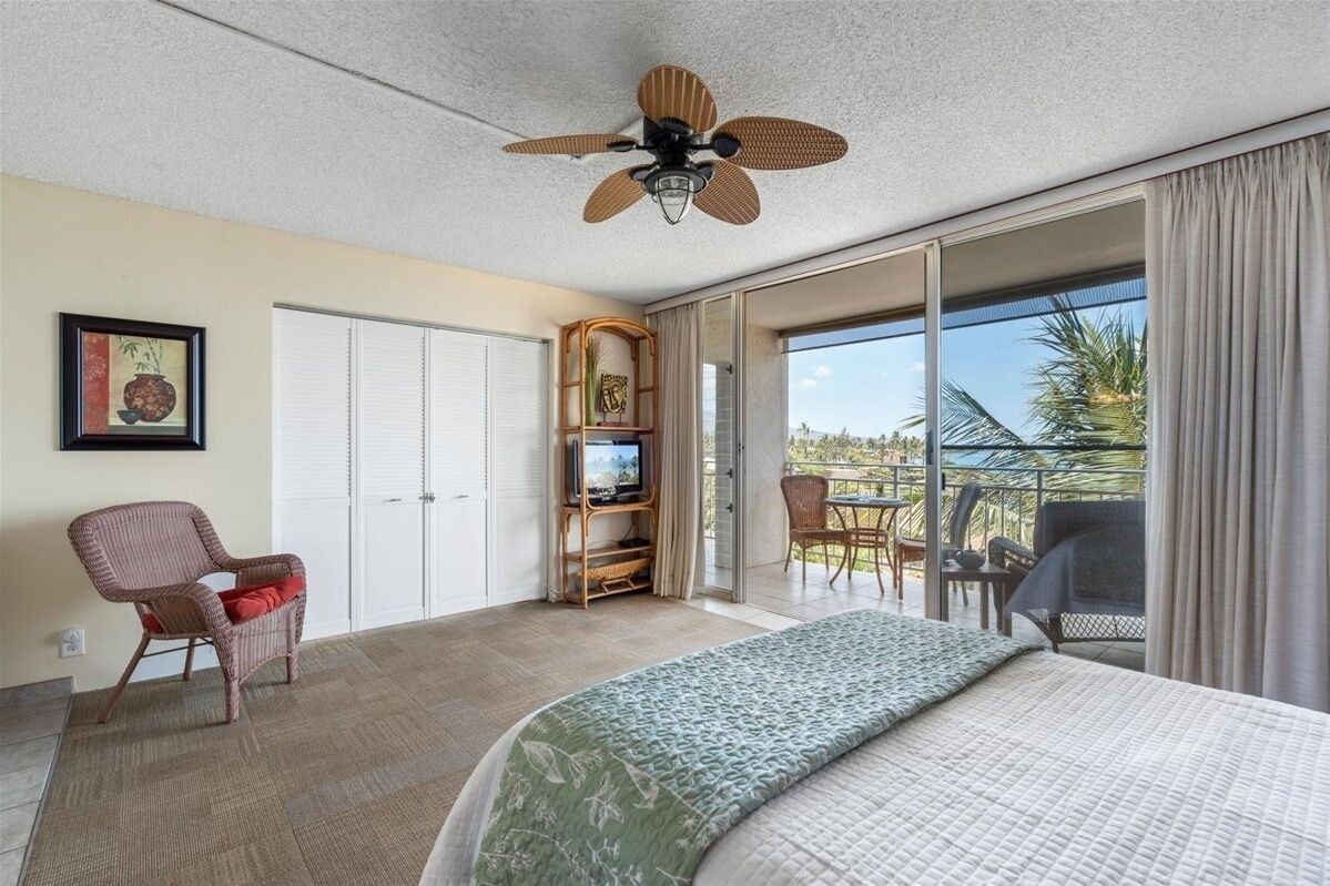 Habitación de hotel con cama, televisión, silla de mimbre, balcón con vistas y ventilador de techo.