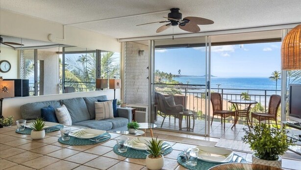 A modern living room with a sofa, dining table, and chairs, a ceiling fan, and a balcony with a view of the ocean. - Island Surf 614 2 Bedroom Apts (Kihei)