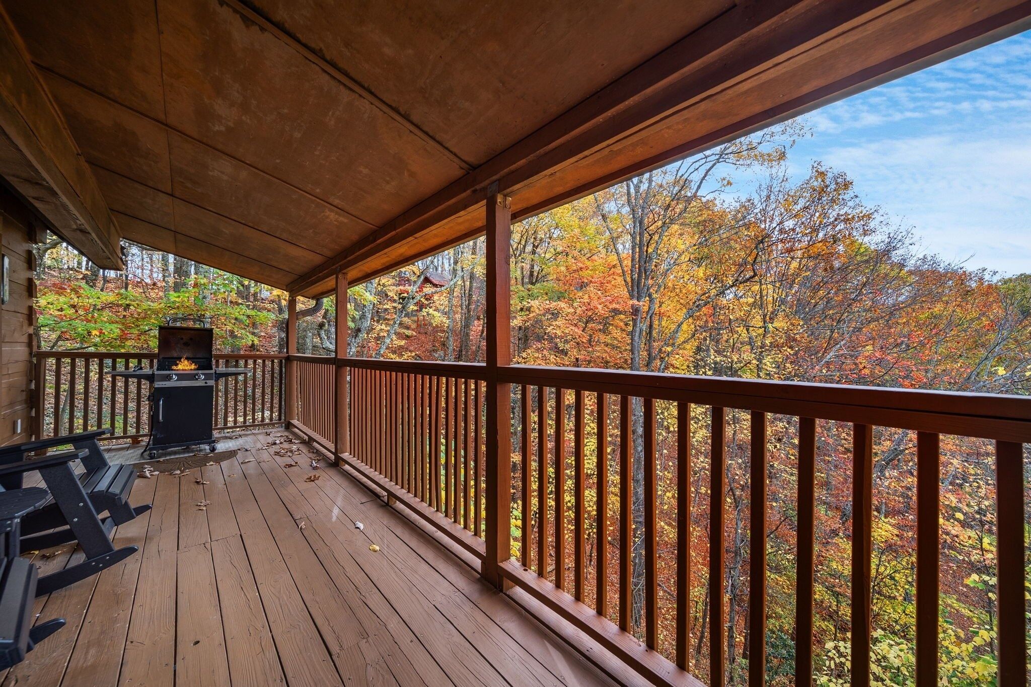 A covered deck with a grill and picnic tables, overlooking a forest with autumn foliage.