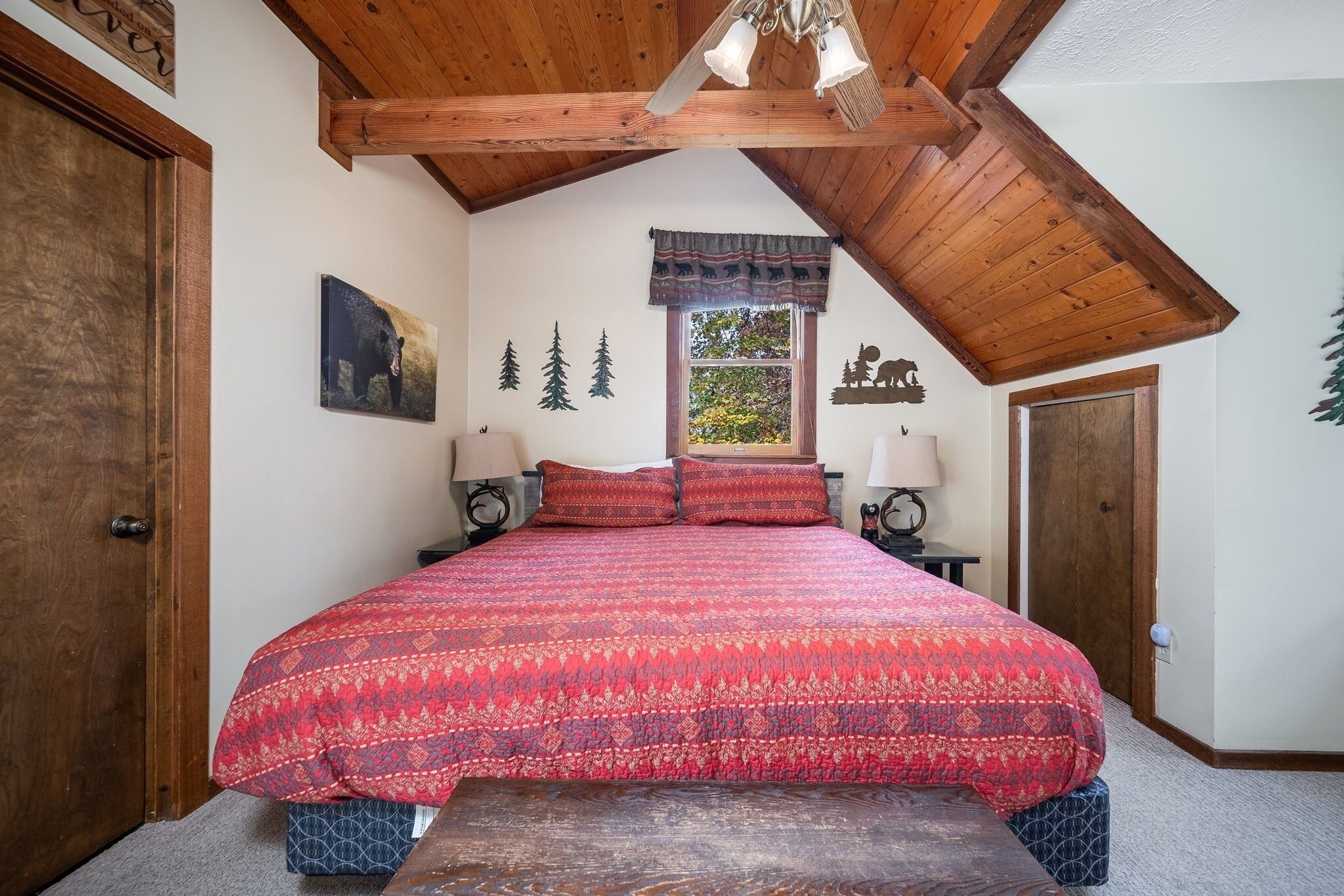 A bedroom with a large bed, wooden ceiling beams, a window with a view, and wall decorations including a bear and trees.