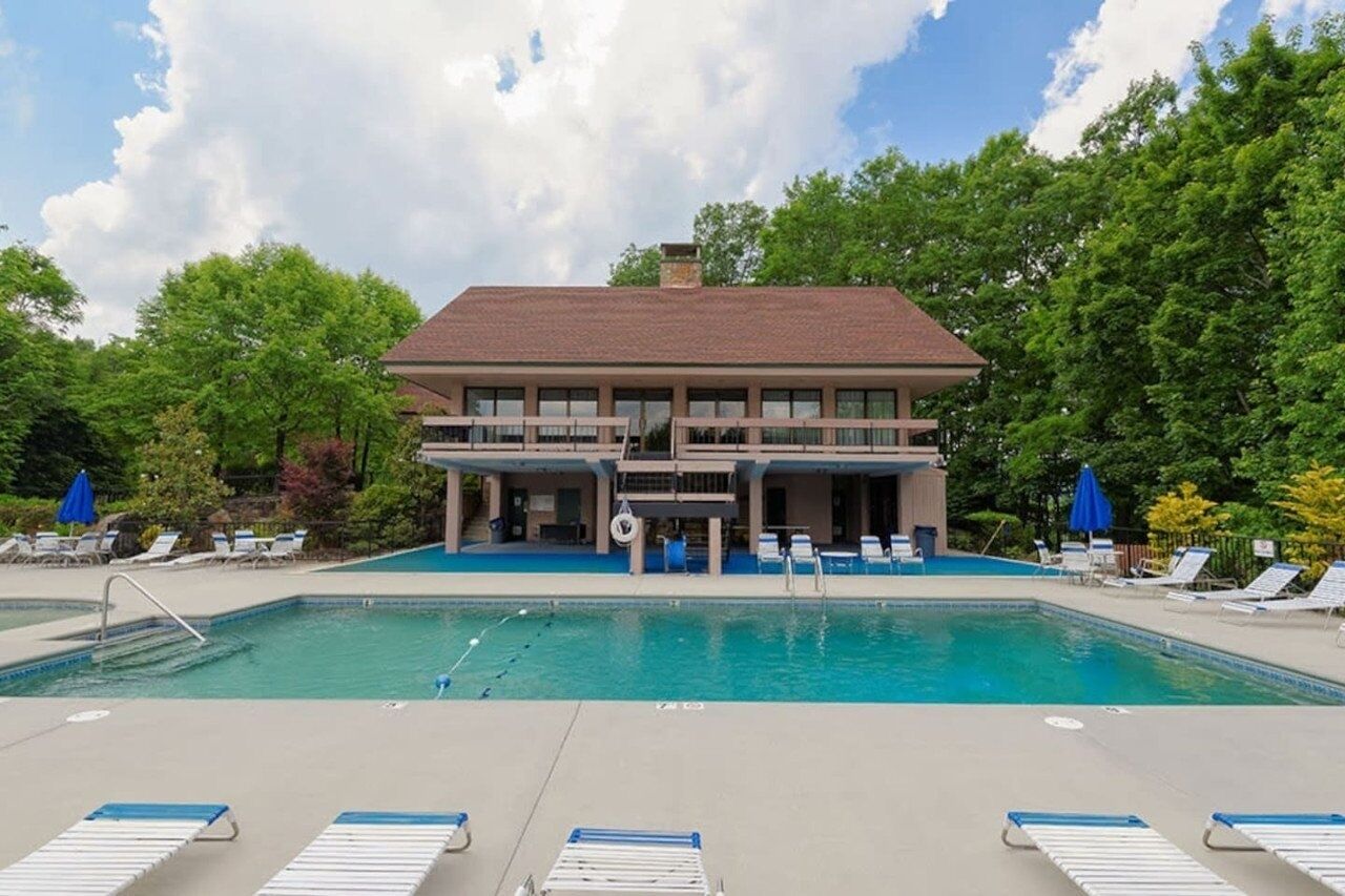 A pool area with lounge chairs and umbrellas, a building with a balcony, and trees in the background.