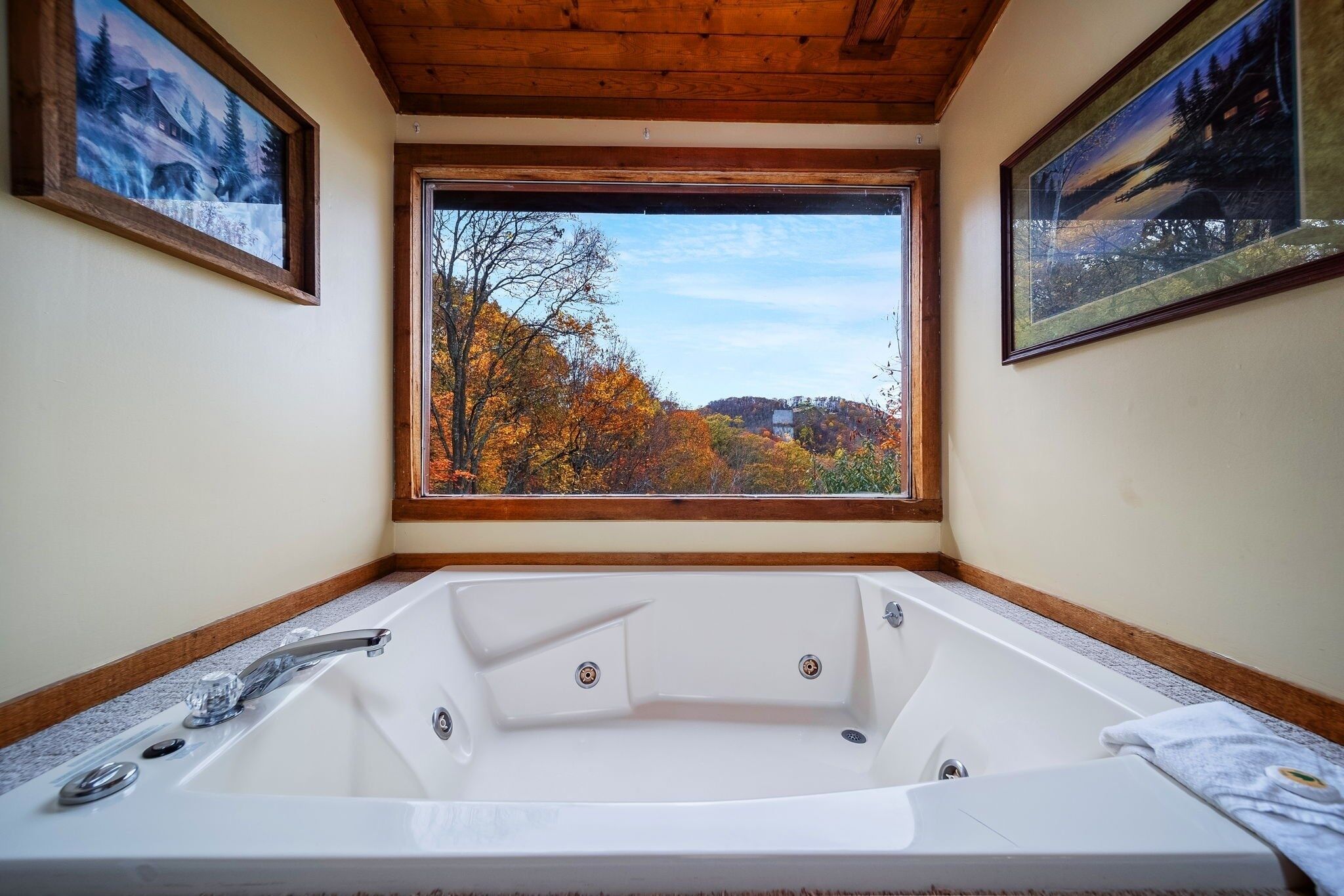 Un baño con una bañera grande, cuadros en las paredes y vistas a árboles otoñales.