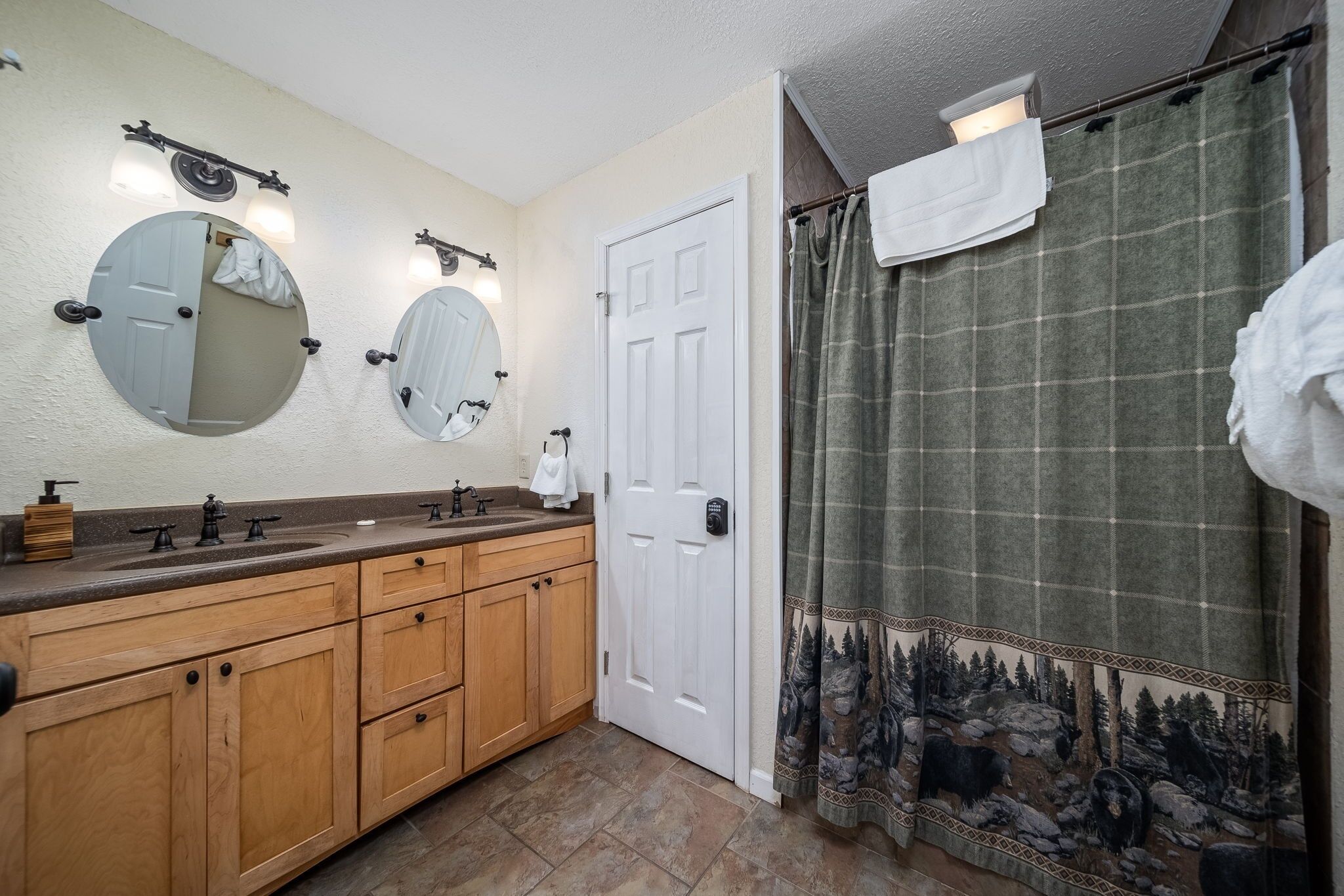 A bathroom with a wooden vanity, two round mirrors, a white door, and a green plaid shower curtain.