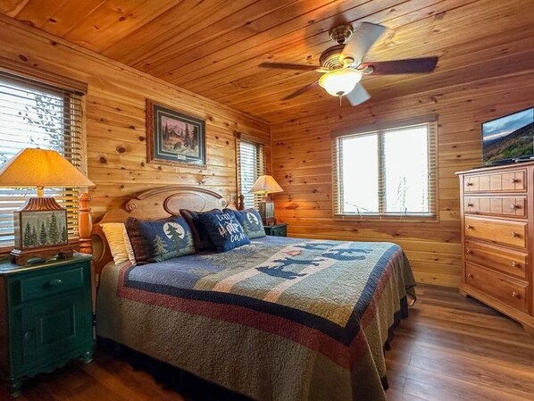 A cozy log cabin bedroom with a bed, wooden dresser, and a ceiling fan.