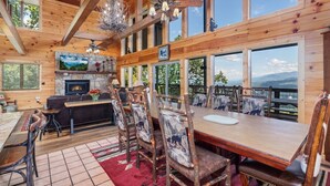 Interior de una cabaña de madera con chimenea, comedor y grandes ventanales que ofrecen vistas a la montaña.