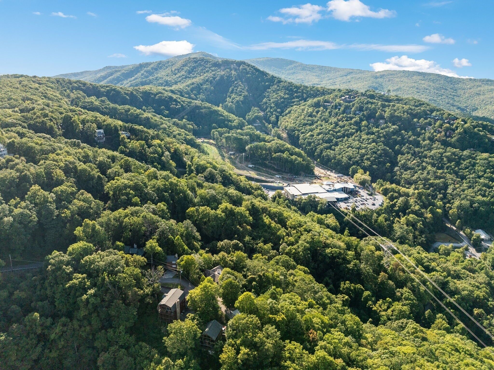 An aerial view of a mountainous area with dense forest, a small building, and a paved area with parked cars.