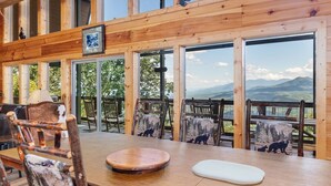 Interior de madera con ventanas amplias, una mesa de comedor, sillas y vistas a montañas.