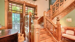 Una majestuosa escalera con detalles tallados, un piano y una guitarra en una habitación cálidamente iluminada, con ventanas amplias y una pared verde.