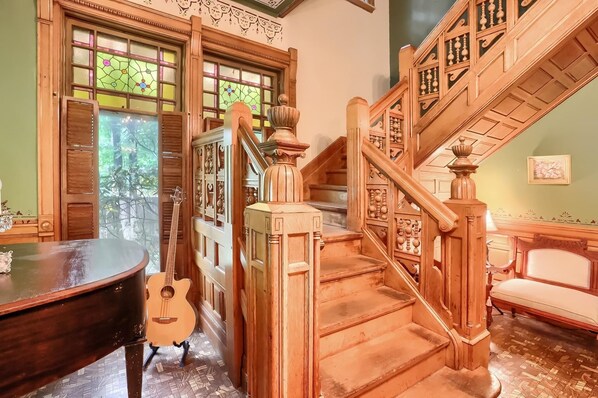 Eine prächtige Treppe mit aufwendigen Holzarbeiten, ein Klavier und eine Gitarre in einem wohlig beleuchteten Raum mit großen Fenstern und einer grünen Wand.
