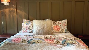 Una cama con ropa de cama floral, un plaid blanco esponjoso y una lámpara con pantalla de cobre.
