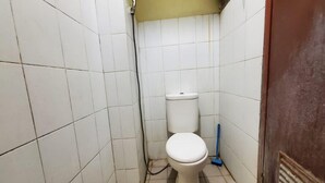 A small, tiled bathroom with a white toilet, a visible pipe, and a broom leaning against the wall.