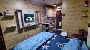 A room with a bed, a desk, and a chair. There is a computer monitor on the wall and a small shelf with a plant.