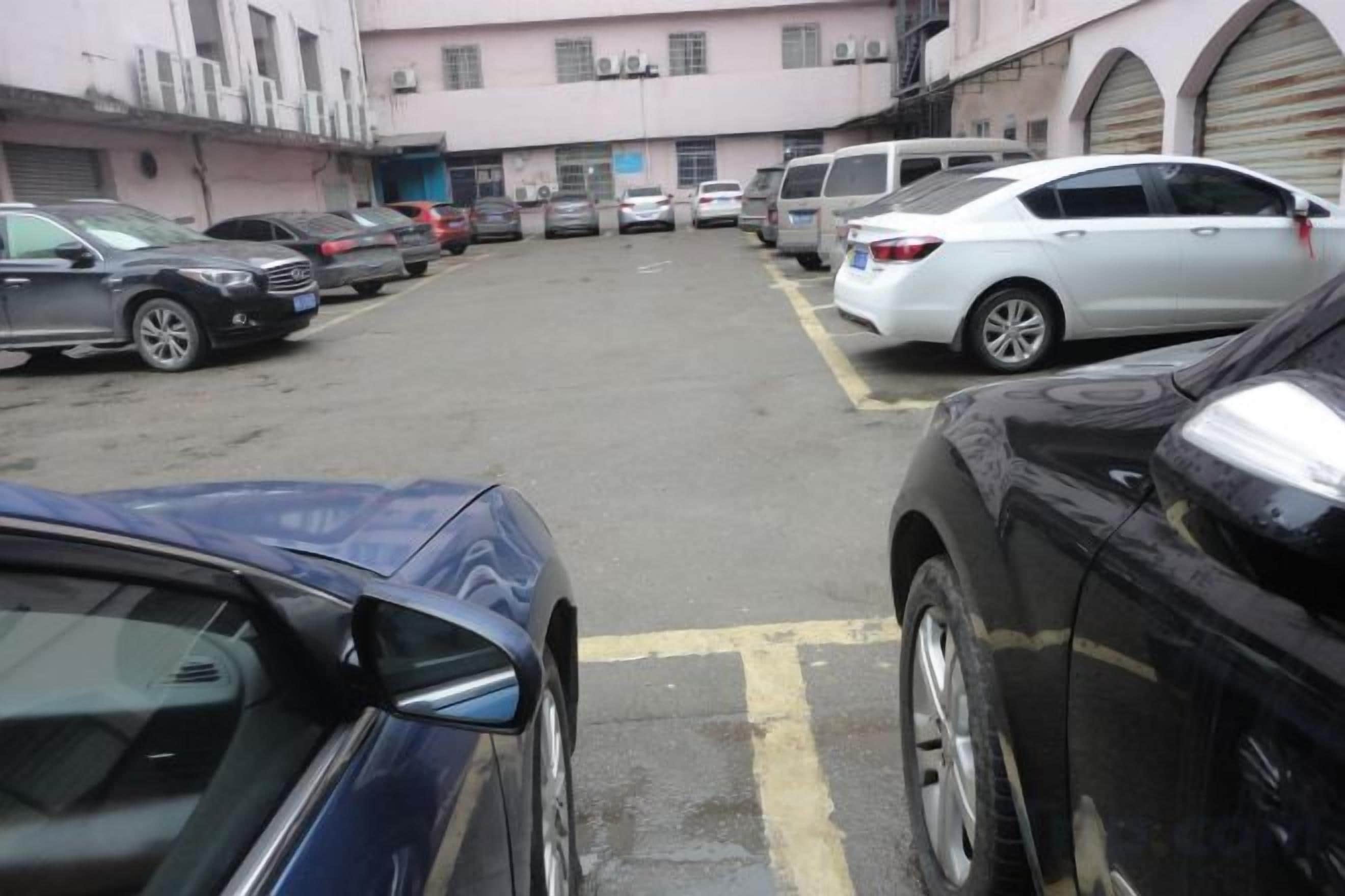 Un estacionamiento con varios coches aparcados en filas, un edificio de fachada rosa al fondo y un cielo despejado.