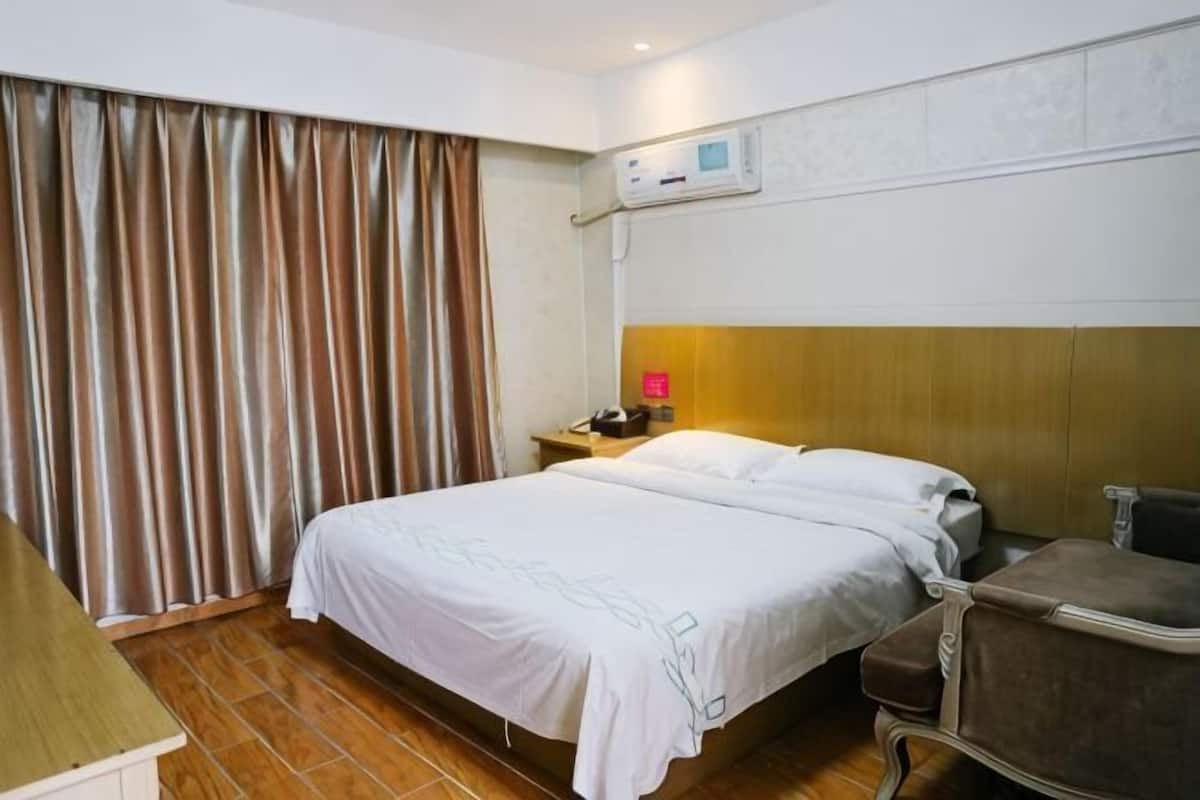 A hotel room with a bed, a bedside table, a nightstand, a lamp, a window with curtains, and a wall-mounted air conditioning unit.