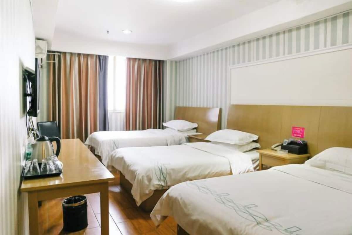 A hotel room with three beds, a wooden table with a kettle, a trash bin, and a nightstand with a phone.