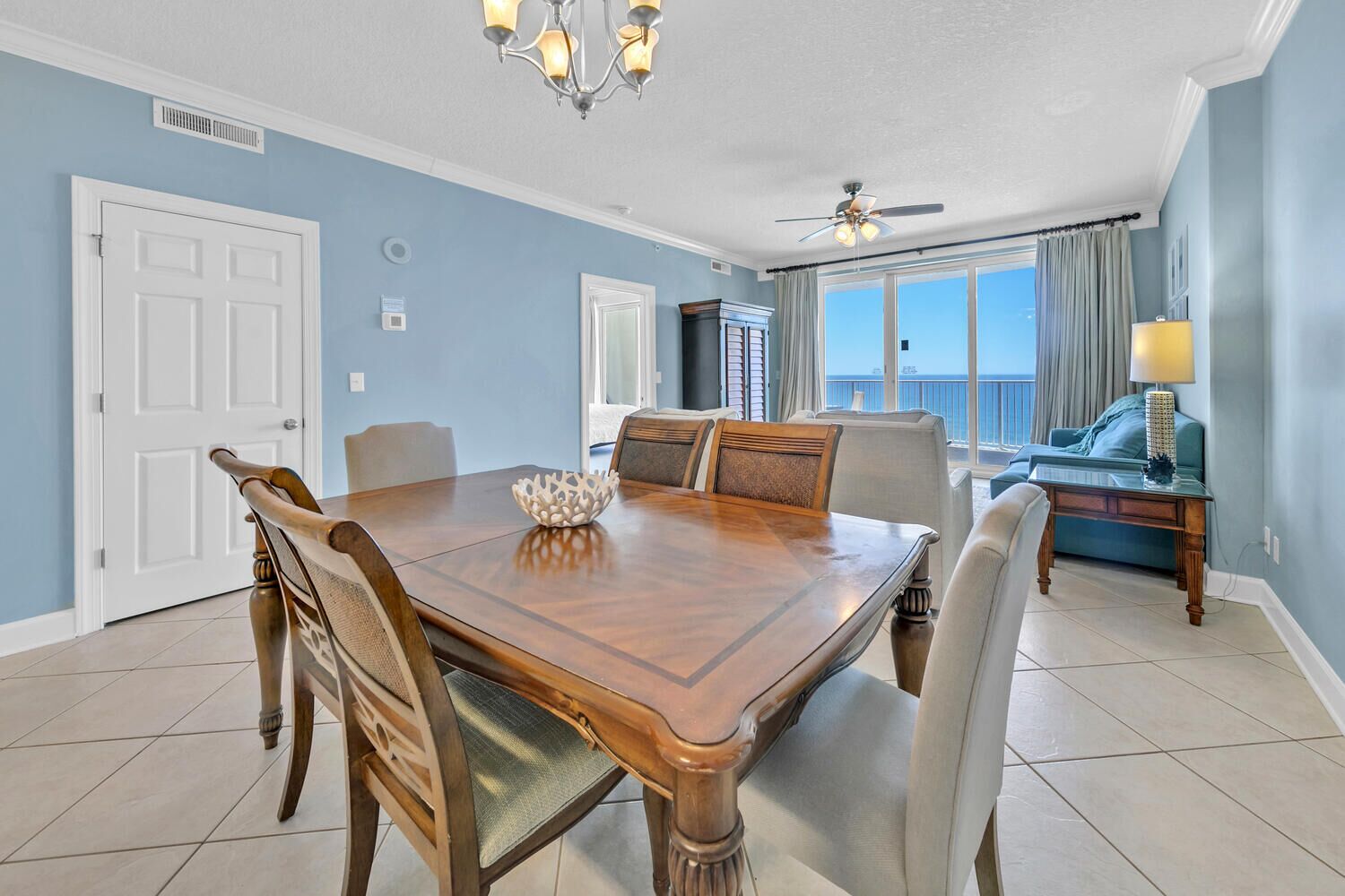 A dining area with a wooden table and chairs, a chandelier, and a view of the ocean through large windows.