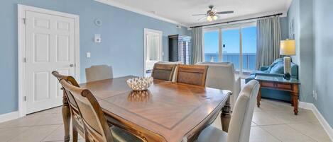 A dining area with a wooden table and chairs, a chandelier, and a view of the ocean through large windows.