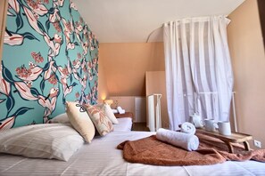 Quarto de hotel com uma cama, almofadas, mesa de cabeceira com abajur, mesinha de madeira com toalhas e uma chaleira, e uma janela com cortinas finas.