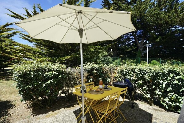 An outdoor dining area with a table and chairs under a large umbrella, surrounded by greenery.