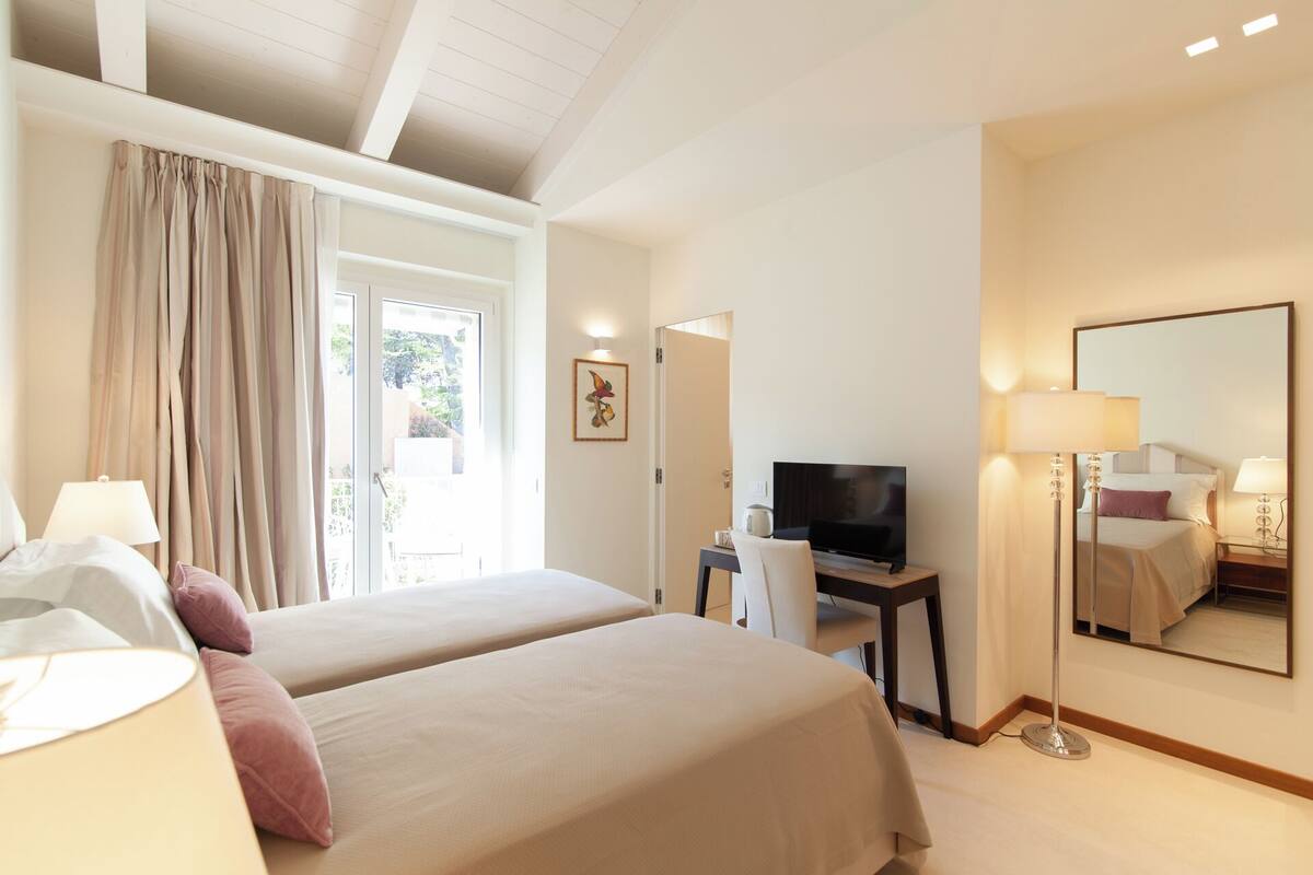 Habitación de hotel con dos camas, un escritorio con televisor, un espejo grande y vistas al exterior a través de una puerta corrediza.