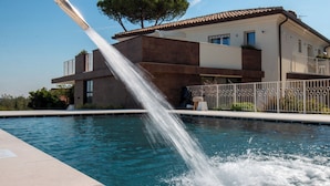 A swimming pool with a water feature, a modern building with a tiled roof, and a tree in the background. - Villa Francesca Relais (Gradara)