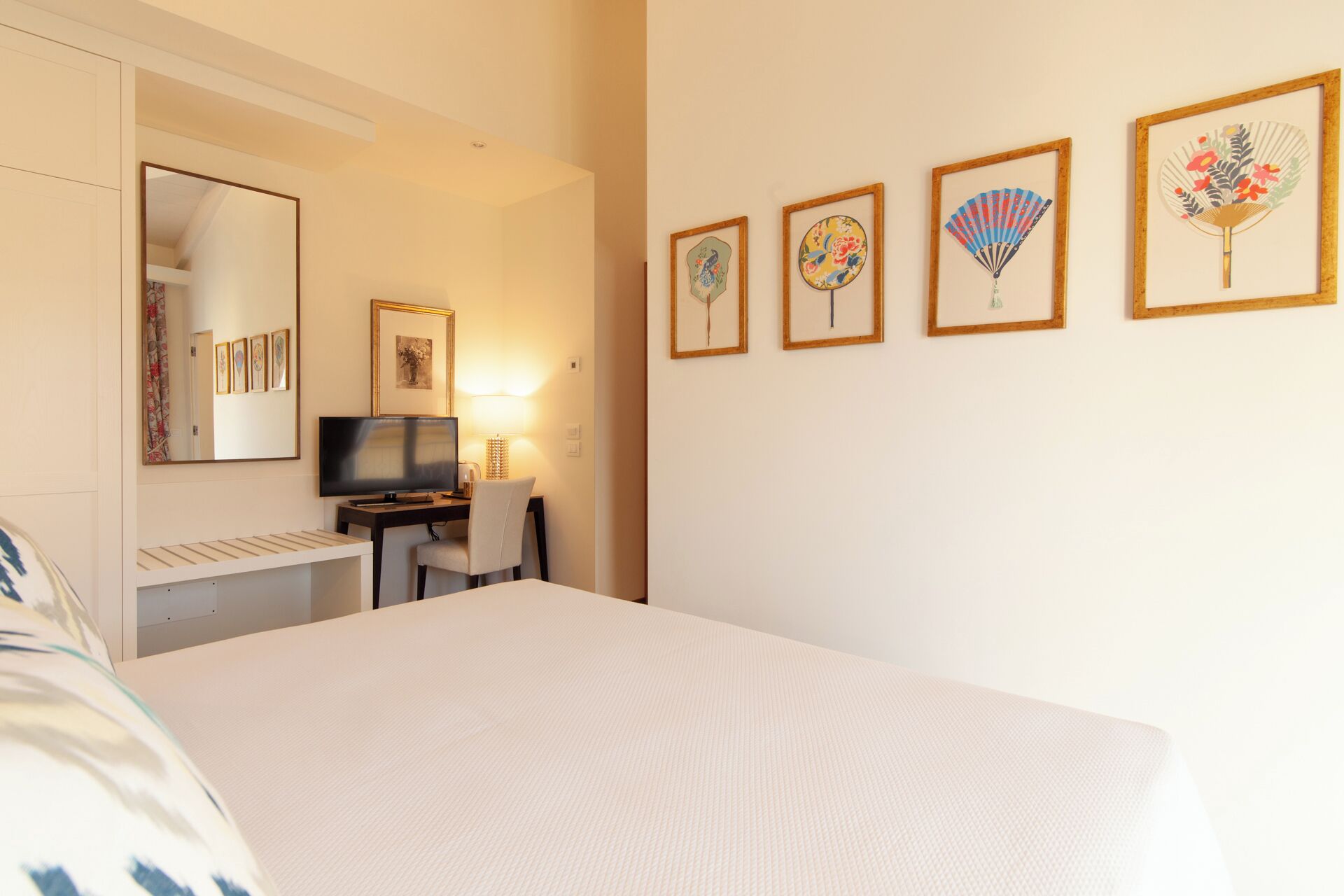A hotel room with a bed, a desk with a computer, a TV, and four framed artworks on the wall.