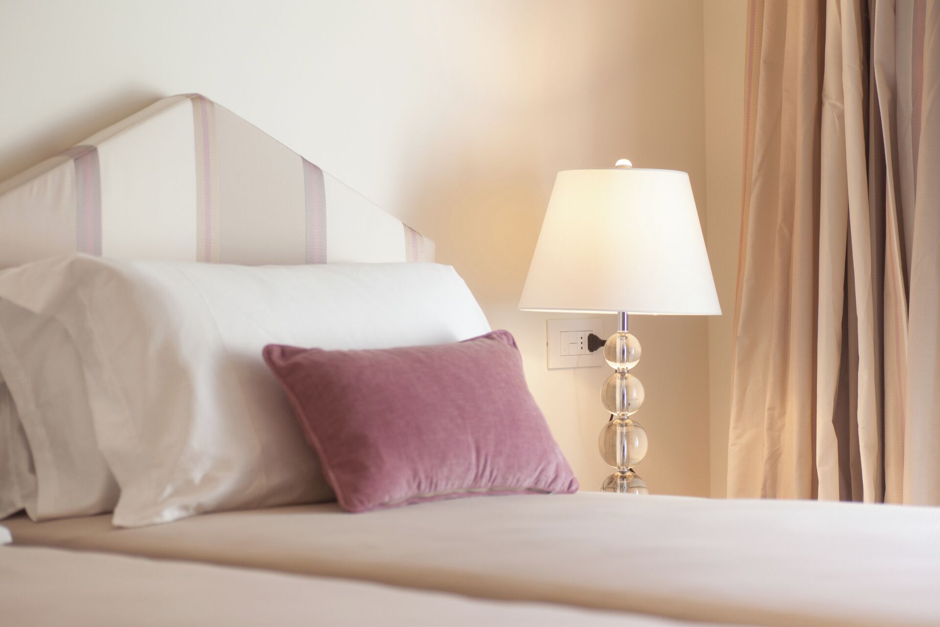Uma cama bem arrumada com roupa de cama branca e roxa, um abajur e uma cortina.