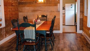 A dining area with a wooden table and chairs, a hanging light fixture, and a kitchen visible through an open door. - DLT Cabins, LLC - Holler Hideaway Cozy house sleeps 4 on 10 Minutes from SNP (Luray)