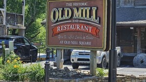 Señal que dice 'The Historic Old Mill and Restaurant', con un muro de piedra y vehículos estacionados cerca.