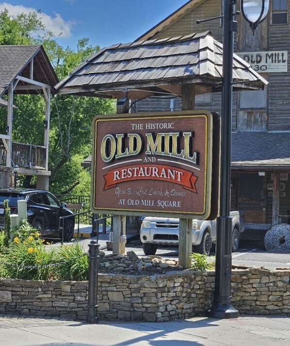 Señal que dice 'The Historic Old Mill and Restaurant', con un muro de piedra y vehículos estacionados cerca.