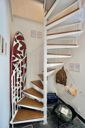 Un escalier en colimaçon, avec une planche de surf appuyée contre le mur, un sac à dos et une valise posés sur le sol.