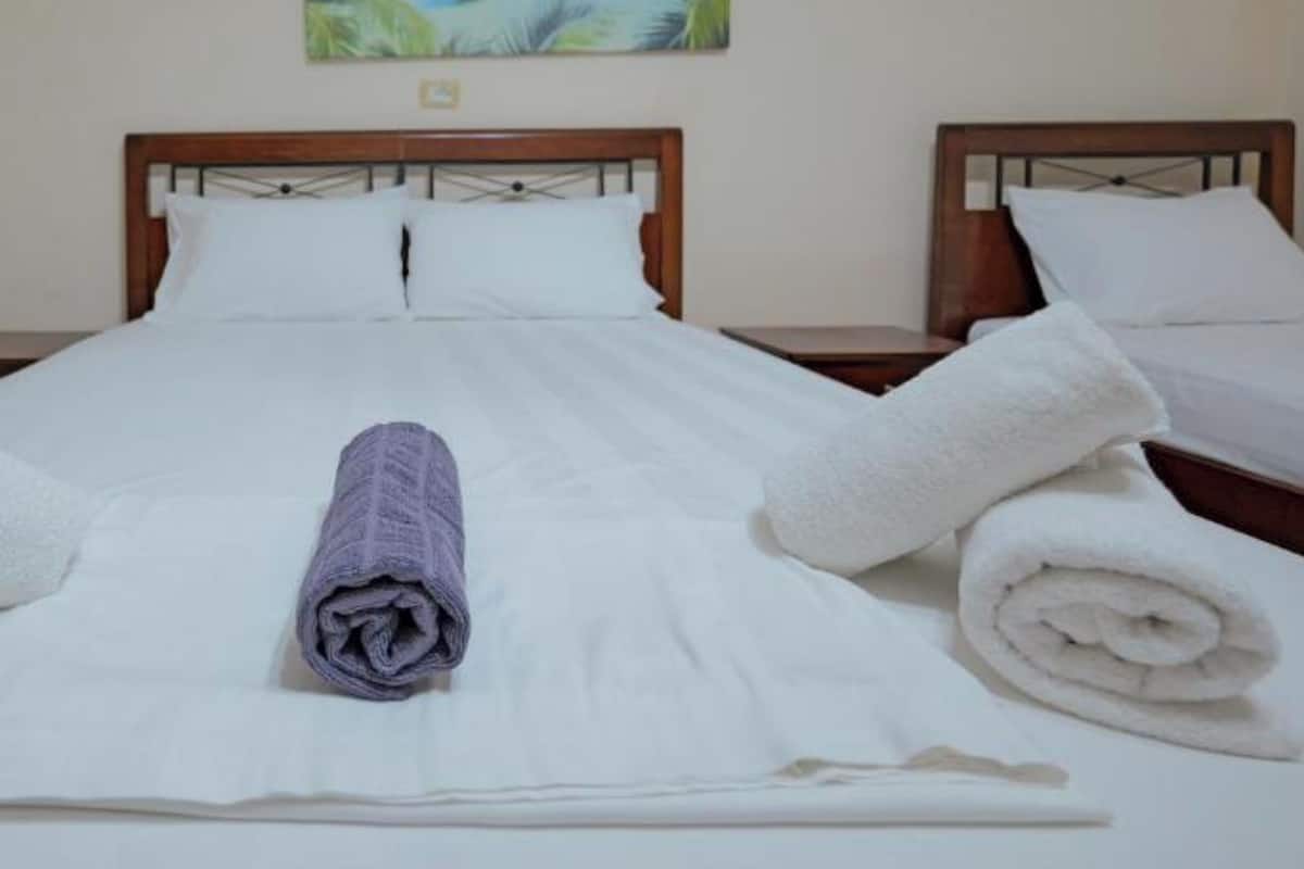 Habitación de hotel con dos camas, ropa de cama blanca, un cuadro de una playa y dos lámparas fijadas a la pared.
