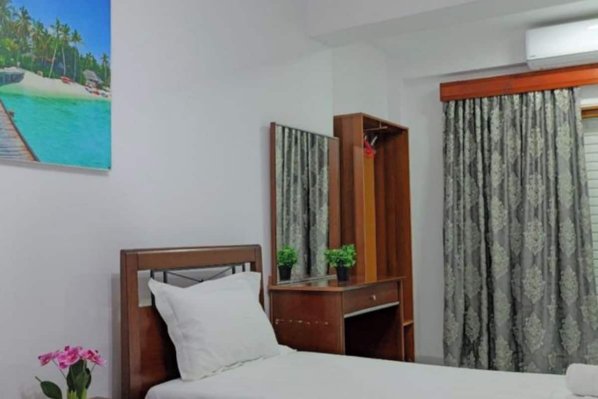 Una habitación de hotel con dos camas, cabecero de madera, una mesita de noche con un jarrón de flores, un escritorio con un espejo y una ventana con cortinas.
