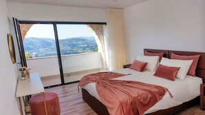 A bedroom with a large bed, a view of the mountains, a small table, and a red ottoman. - Vista D'Ouro- Luxury Villa in the mountains (Lavradas, Ponte da Barca)