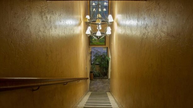 A narrow, wooden-floored hallway with a chandelier and a view of greenery through a window at the end. - Hotel Dikran (Los Angeles)