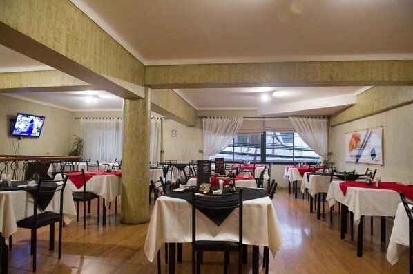 Un restaurante con varias mesas, algunas con manteles rojos, y un televisor montado en la pared.