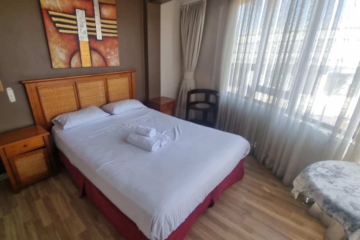 A hotel room with a large bed, wooden headboard, and a view of the outdoors through sheer curtains.