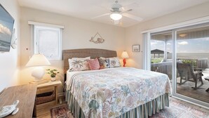 Ein Schlafzimmer mit einem großen Bett, einem Deckenventilator, einem Nachttisch mit Lampe, einem Holztisch und Meerblick durch eine Schiebeglastür.
