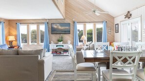 Un vaste salon doté d’un haut plafond, de murs en bois, d’une grande fenêtre avec des rideaux, d’un ventilateur de plafond, d’un téléviseur et d’un coin repas avec des meubles blancs.